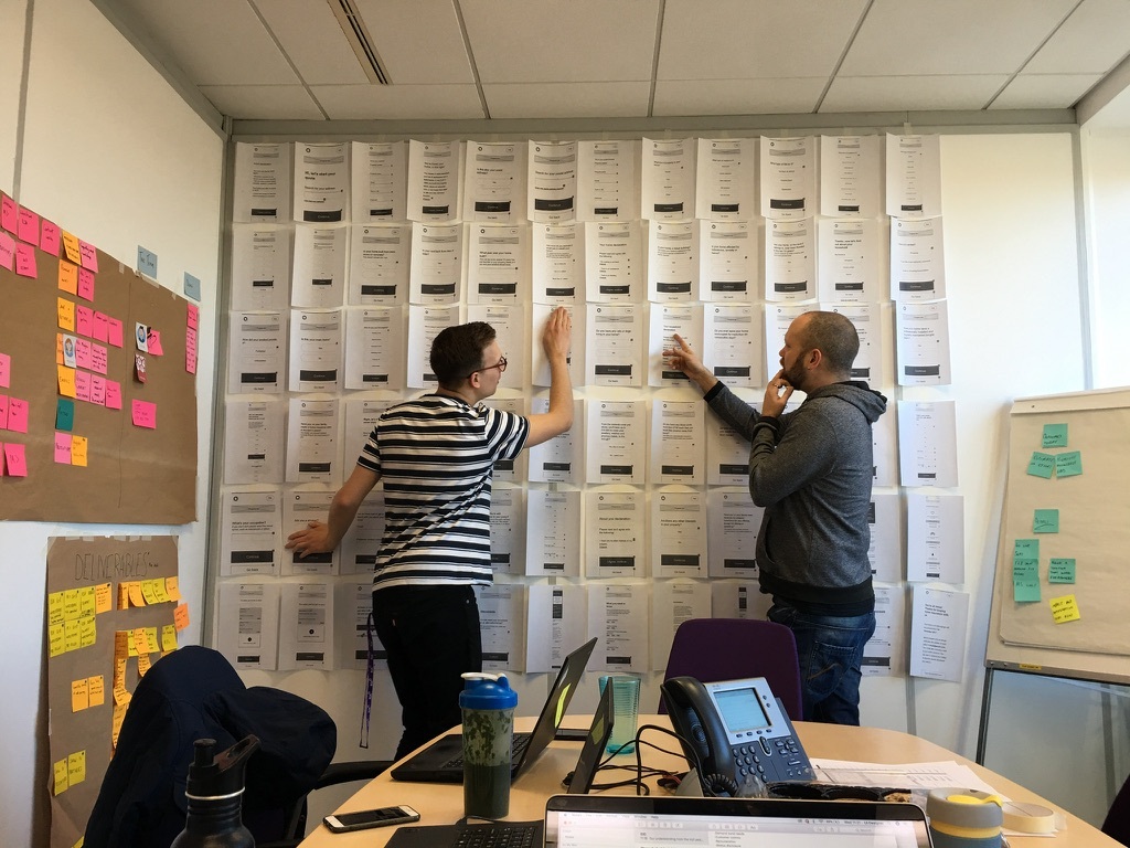 Ben Hills-Jones and I looking at a wall covered in A4 sheets of paper. Each sheet has a screenshot from the prototype.